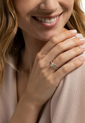 Diamanten verlovingsring met een ronde centrale steen, gezet in een slanke zilveren band met subtiele accentstenen. Hand rustend op een roze stof.
