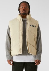 Chaleco acolchado beige con cuello alto, patrón acolchado, bolsillos laterales y un parche de logo negro, usado sobre un suéter gris y una camisa de capas.