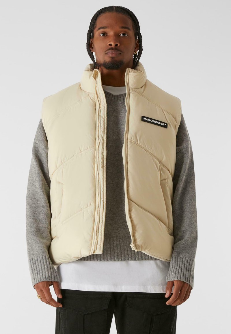 Chaleco acolchado beige con cuello alto, patrón acolchado, bolsillos laterales y un parche de logo negro, usado sobre un suéter gris y una camisa de capas.
