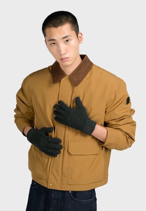 Bruine bomberjack met een corduroy kraag en zwarte handschoenen. De handschoenen hebben een logo patch. Het model draagt donkere jeans.