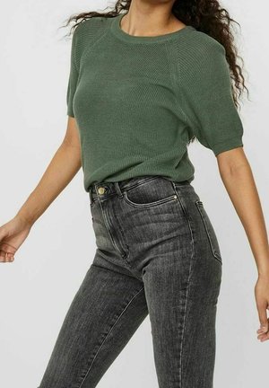 Osoba ubrana w zielony krótki sweter z dzianiny i wysokiej talii wyblakłe czarne obcisłe dżinsy na prostym tle.