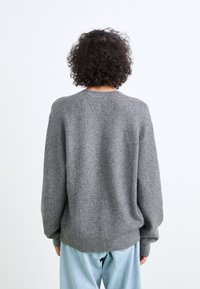 Maglione grigio in lana con scollatura rotonda, spalle scese e vestibilità rilassata. Tessuto morbido e orlo a coste. Indossato con jeans azzurri chiari.