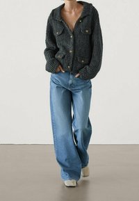 Cardigan grigio scuro lavorato a maglia grossa con colletto, bottoni dorati e due tasche frontali, abbinato a jeans a gamba larga azzurri e scarpe beige.