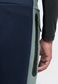 Ναυτικό και ανοιχτό πράσινο ύφασμα, με εφαρμοστό σχέδιο. Περιέχει κάθετη τσέπη με φερμουάρ σε μαύρο χρώμα. Ορατή λεπτομέρεια ραφής.