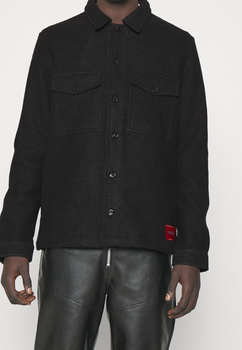 Homme portant une veste noire boutonnée avec deux poches poitrine et un pantalon en cuir noir avec une fermeture éclair à l’avant.