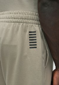 Gaismas bēša krāsas sporta bikses ar elastīgu vidukļa jostu un sānu kabatu, kurā ir septiņas melnas horizontālas svītras kā dizaina elements.