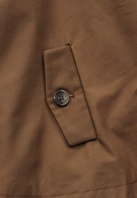 Καφέ ύφασμα με ορθογώνια τσέπη με καπάκι, ασφαλισμένη με σκούρο στρογγυλό κουμπί, δείχνοντας την υφή του υφάσματος και λεπτομέρειες ραφής.
