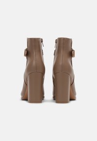 Un par de botas tobilleras en taupe con tacones en bloque y cremalleras laterales, vistas desde atrás sobre un fondo blanco.