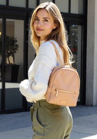 Zaino in ecopelle rosa chiaro con dettagli in zip dorati, di dimensioni piccole e forma arrotondata. Il modello indossa un maglione bianco e pantaloni verde oliva.