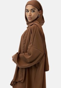 Brun, flödig hijab och abaya-set tillverkat av mjukt, lättviktigt tyg. Har lösa ärmar och lager på lager drapering för komfort och blygsamhet.