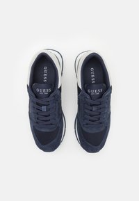 Sneaker blu scuro realizzati in materiali scamosciati e in rete, con suola bianca, lacci rotondi e il marchio "GUESS EST. 1981" sulla linguetta.