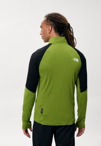 Zielona i czarna bluzka z długim rękawem z wysokim kołnierzem, bezszwowymi ramionami, teksturowanym materiałem i logo na plecach.