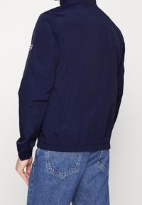 Lehká tmavě modrá bunda s vysokým límcem, elastickým lemem a jemným leskem. Na levém rukávu je logo a dlouhé rukávy.