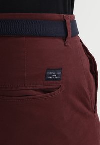 Burgundyröda tygshorts med ett marinblått vävt skärp, sidoficka och en svart etikett med vit text som visar varumärkesinformation och skötselföreskrifter.