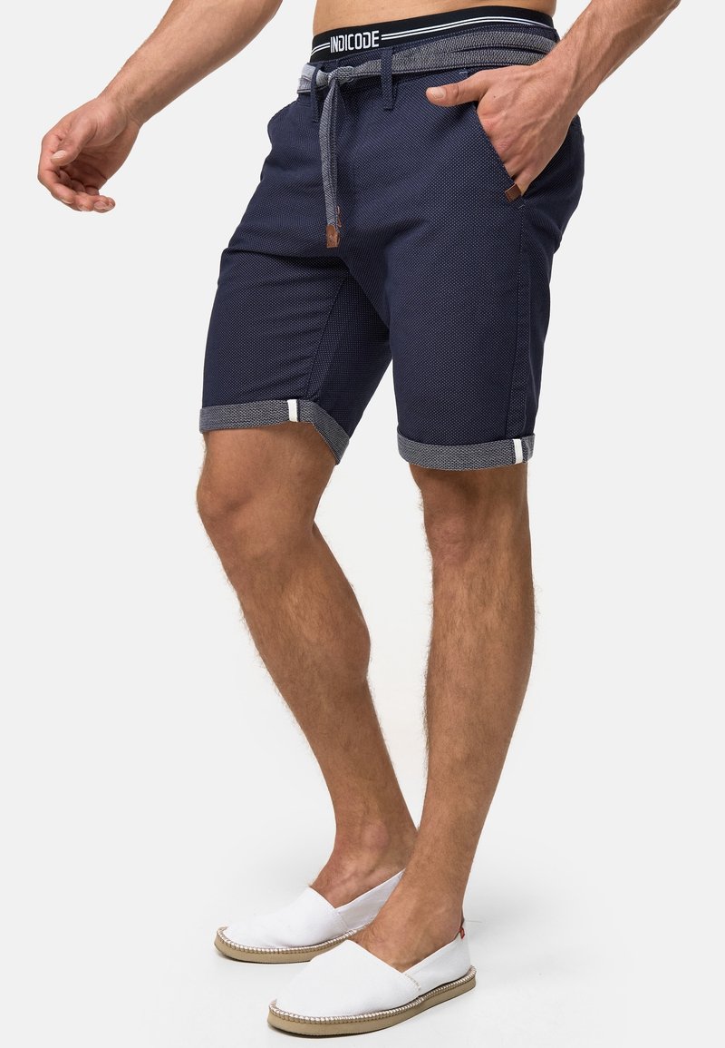 Homme portant un short texturé bleu marine avec des ourlets retroussés, une ceinture en corde et des chaussures blanches sans lacets, debout devant un fond uni.