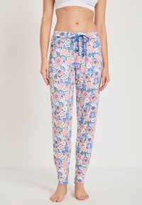 Vrouw in blauwe bloemen drawstring pants en een witte cropped top, staand op blote voeten tegen een effen achtergrond.