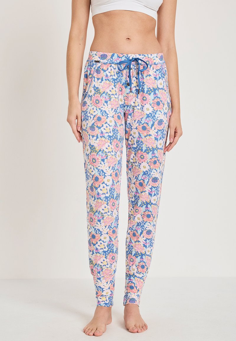 Vrouw in blauwe bloemen drawstring pants en een witte cropped top, staand op blote voeten tegen een effen achtergrond.