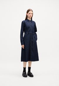 Jeansklänningskjol i mörkblått. Har en knappslutning framtill, långa ärmar, krage, midjebälte och sidofickor. Platta svarta skor.