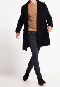 Cappotto di lana navy sopra un maglione lavorato a maglia color beige, abbinato a pantaloni grigi scuri e stivaletti neri, caratterizzato da una silhouette pulita e dettagli minimi.
