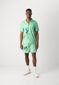 Grøn kortærmet skjorte og matchende shorts med et blomsterdesign i gul og blå. Lavet af letvægtsstof med en afslappet pasform.