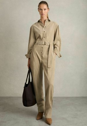 Beige utility overall med krave, knaplukning, bælte i taljen og lommer; parret med en sort tote bag og brune hæle.