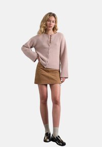 Femme debout, main sur la hanche, portant un pull rose boutonné, une mini-jupe en suède marron, des chaussettes beiges et des mocassins noirs à motifs.