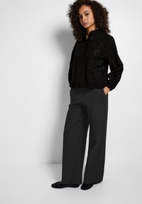 Blusa nera ricamata con maniche lunghe e orlo a scallop, abbinata a pantaloni larghi grigi e ballerine nere, con una vestibilità comoda.
