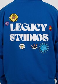 Blauwe sweatshirt met grote witte tekst "LEGACY STUDIOS" en kleurrijke borduursels van bloemen en zon. Zachte stof, casual pasvorm.