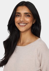 Helles beiges Sweatshirt mit weitem Ausschnitt, gerippter Textur und weicher Stoffqualität. Langes, dunkles Haar umrahmt das Gesicht, das ein sanftes Lächeln zeigt.