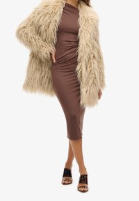 Fuskpäls cremfärgad kappa över en figursydd brun midiklänning. Öppen tå svart leopardmönstrad glidsandal. Släta texturer och lagerdesign.