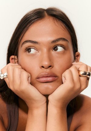 Deux anneaux, un en argent et un en or, portés à des doigts pendant que la personne repose son menton sur ses mains. Peau lisse et maquillage naturel.