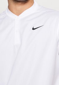 Bílá polokošile s límečkem, vyrobená z hladké látky, se zapínáním na tři knoflíky a malým černým logem Nike na levé straně hrudi.