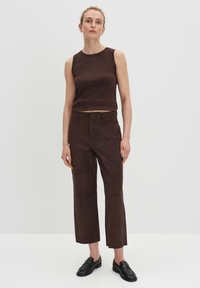 Braunes ärmelloses Oberteil und braune hochtaillierte Hose mit glatter Textur. Flache schwarze Schuhe ergänzen das Outfit. Einfaches, körpernahes Design.