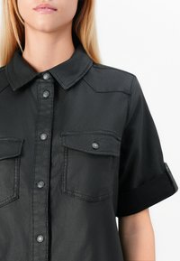 Maglietta nera a maniche corte realizzata in un materiale leggero e lucido. Presenta una chiusura con bottoni, due tasche sul petto e risvolti sui polsini.