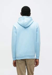 Gaiši zils kapuces džemperis ar mīkstu faktūru, ribotas aproces un brīvu piegriezumu. Aizmugures skats atklāj gludas šuves un regulējamu kapuci.