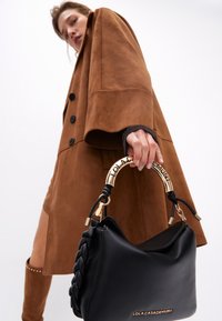 Una modella in un cappotto marrone tiene una borsa nera con una tracolla intrecciata. L'attenzione è sugli accessori e sull'outfit alla moda.