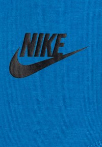Sininen kangas teksturoidulla neuleella. Musta Nike-logo, jossa on näkyvä swosh-muotoilu. Ei muita korostuksia tai kuvioita näkyvissä.