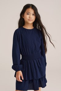 Tengerészkék színű gyermekruha hosszú ujjakkal, textil hatású anyaggal, rakott derékkal és több rétegből álló szoknyával. Finom mintával és kerek nyakkivágással rendelkezik.