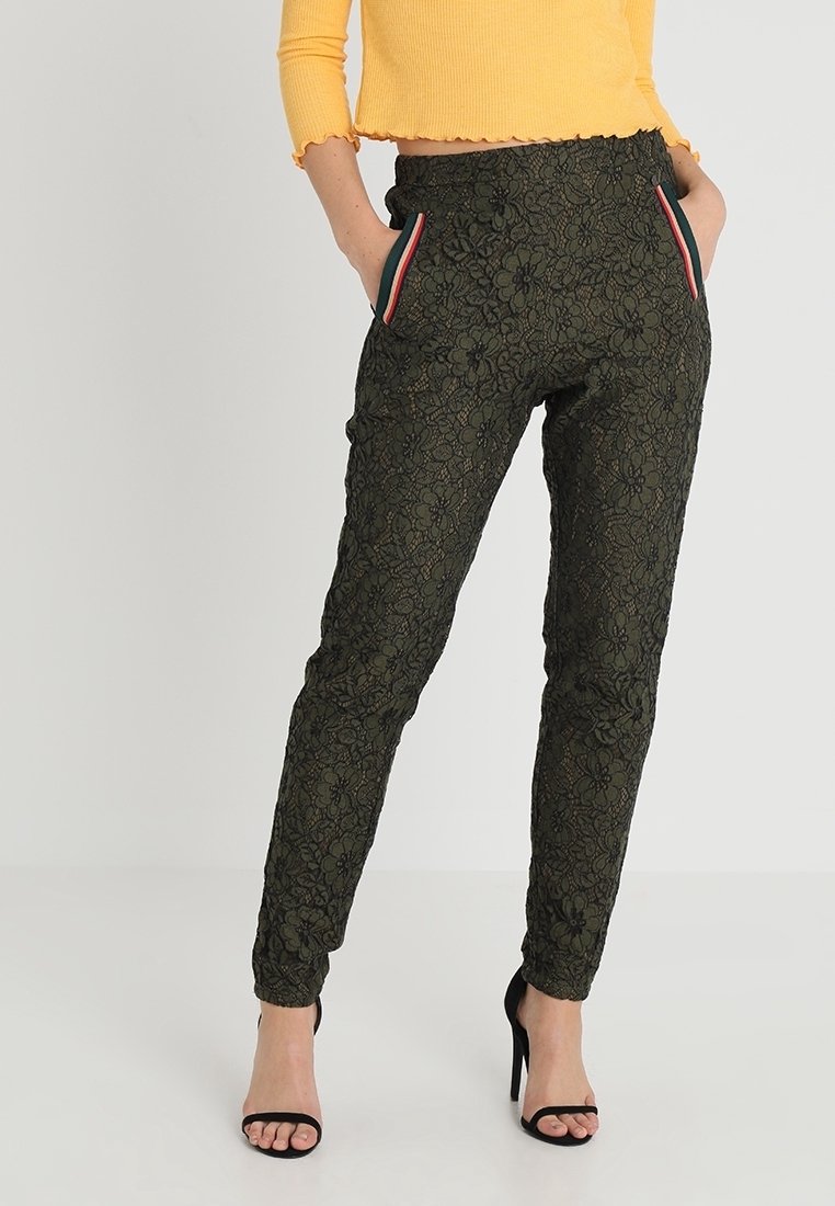 Femme portant un pantalon noir en dentelle florale texturée avec bordure rayée aux poches, associé à un pull court jaune et des talons ouverts noirs.