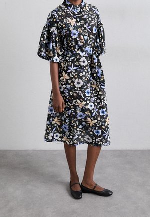 Femme portant une robe noire fleurie mi-longue aux genoux avec des manches bouffantes et une ceinture à la taille, associée à des chaussures plates Mary Jane noires.
