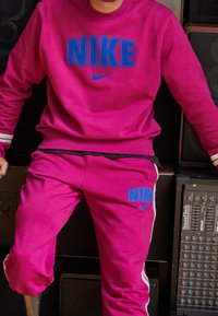 Felpa Nike e pantaloni della tuta in un vivace rosa con logo blu. La felpa ha un colletto a girocollo, i pantaloni presentano strisce laterali e branding.
