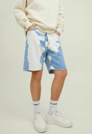 Person trägt weiße und blaue Batik-Denim-Shorts mit den Händen in den Taschen, weiße Sneakersocken, weiße Sneakers und einen beigen Pullover.