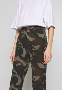 Personne portant un t-shirt blanc ample rentré dans un pantalon camouflage vert et marron taille haute, debout devant un fond uni.