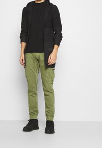 Felpa nera con struttura a coste, abbinata a pantaloni cargo verde oliva con tasche. Calzature nere e strutturate. Design semplice e funzionale.