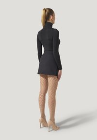 Kvinna med blont hästsvans som bär en svart långärmad polotröja, kort svart kjol, beige prickiga strumpbyxor och nude högklackade skor, stående med sidan vänd.