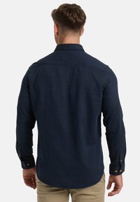 Mann mit kurzen braunen Haaren, der ein langärmeliges dunkelnavyblaues Hemd und beigen Hosen trägt, von hinten vor einem schlichten weißen Hintergrund dargestellt.