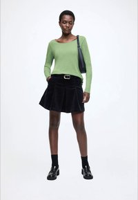 Zelený top s dlouhým rukávem ve spojení s černou plisovanou sukni z manšestru, černou masivní obuví a malou černou kabelkou přes rameno. Hladká textura.