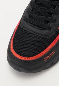 Nærbilde av svart og rød Kappa-sneaker tå med mesh-stoff, svarte lisser og logo på siden mot en hvit bakgrunn.