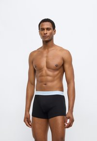 Μαύρα boxer εσώρουχα με ζώνη στη μέση σε γαλάζιο χρώμα της Calvin Klein, με εφαρμοστή σχεδίαση και λεία υφή. Το μοντέλο παρουσιάζει ουδέτερη στάση.
