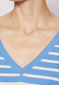 Blauwe en witte gestreepte v-hals top gemaakt van zachte stof, met een smalle halslijn en stikdetails, en een klein logo aan de zijkant.